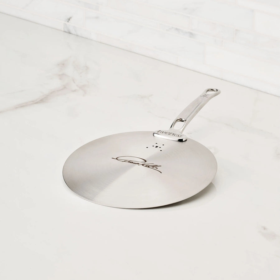 The Thomas Keller Insignia Commercial Clad Stainless Steel Universal Lid, with handle, steam vents, and engraved signature, sits on a white marble countertop—an ideal addition to eco-friendly cookware.