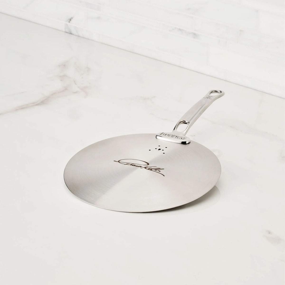 The Thomas Keller Commercial Clad Stainless Steel Universal Lid, featuring an engraved handle, sits on a white marble countertop—an ideal addition to your eco-friendly cookware collection.