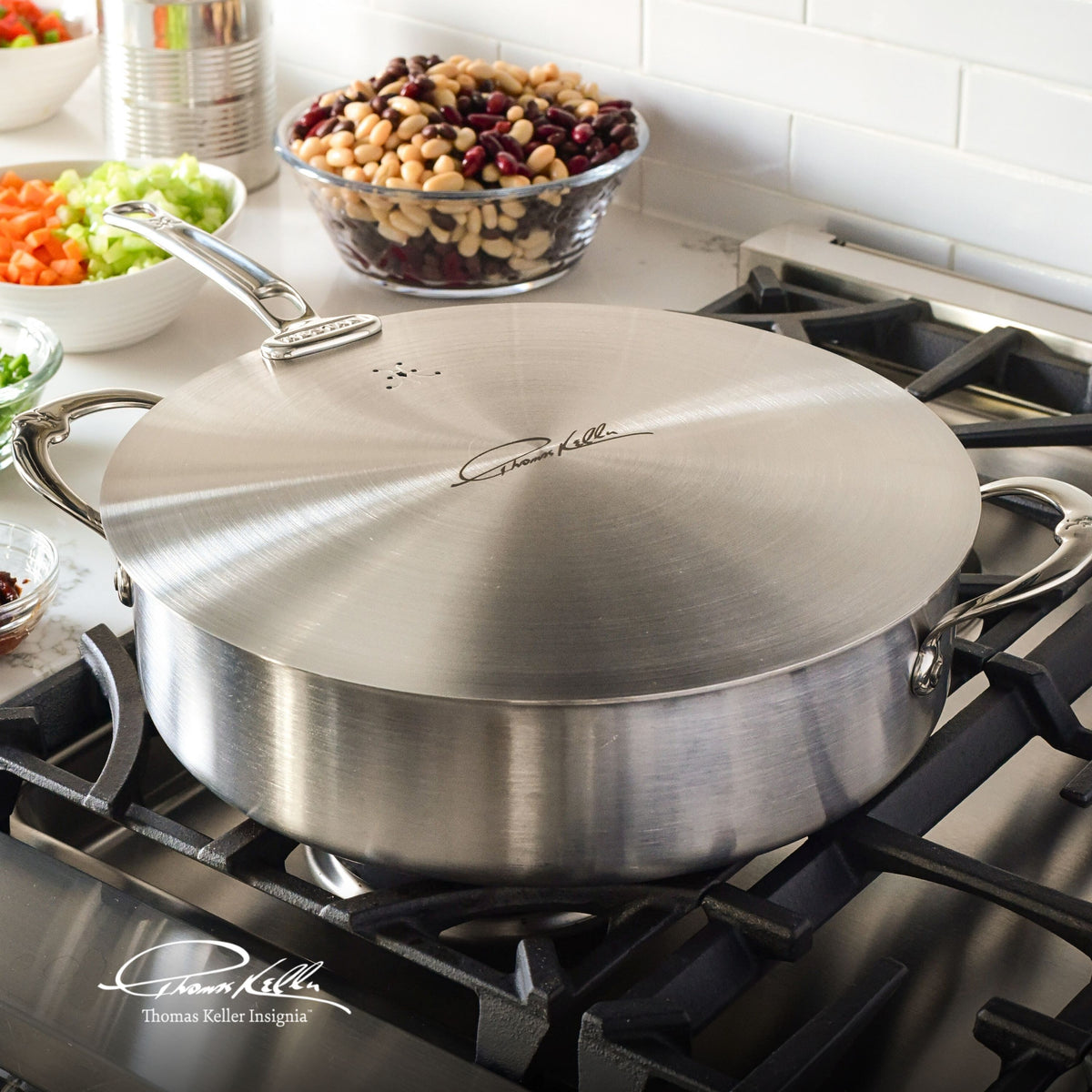 A Thomas Keller Commercial Clad stainless steel pot with a Stainless Steel Universal Lid, featuring a signature engraving, sits on a gas stove. Bowls of chopped vegetables and beans are on the white countertop in the background.