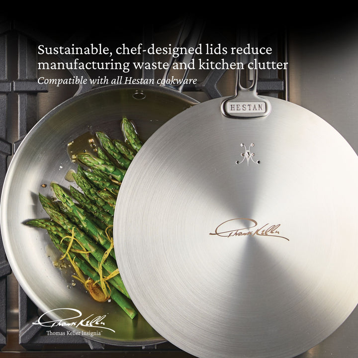 A stainless steel pan with asparagus is on a stovetop next to a Thomas Keller Insignia Commercial Clad Stainless Steel Universal Lid, promoting eco-friendliness and compatibility with all Hestan cookware.