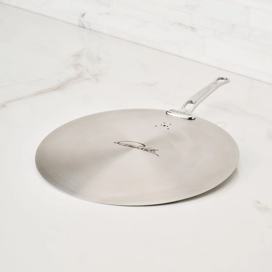 A Thomas Keller Insignia Commercial Clad Stainless Steel Universal Lid with a handle rests flat on a white marble countertop, featuring an eco-friendly design, a logo, and three small holes in a triangle near the handle.