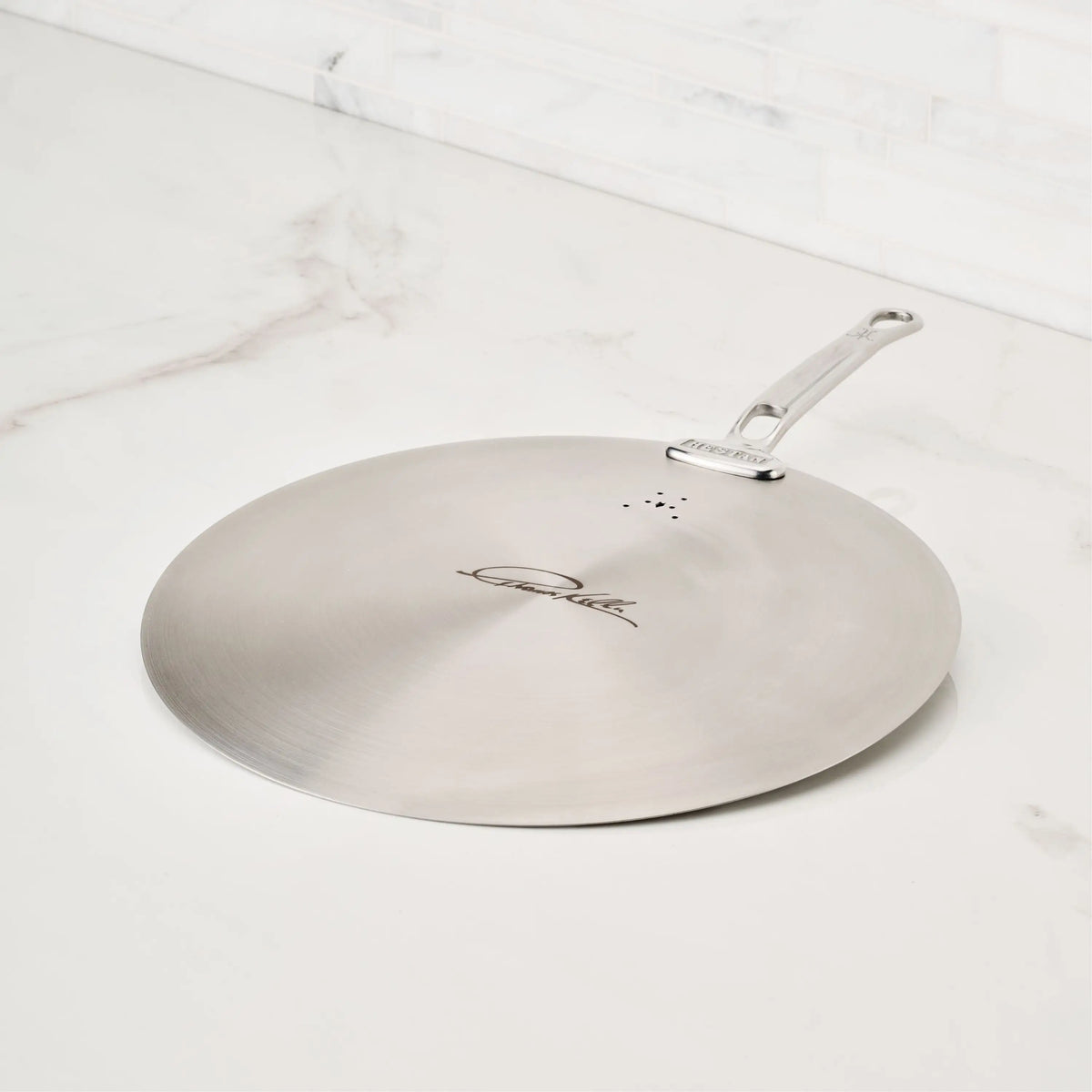 A Thomas Keller Commercial Clad Stainless Steel Universal Lid with handle rests on a white marble countertop. Made to pair with eco-friendly cookware, it features small vent holes and a center signature engraving.