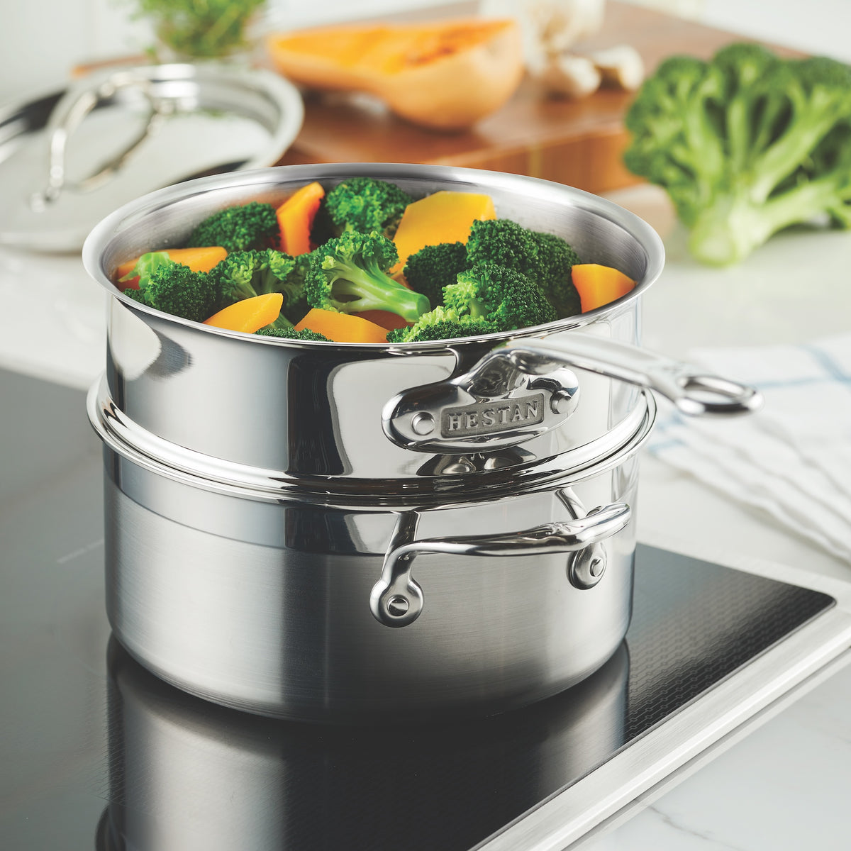 Using the Hestan Provisions Stainless Steel Steamer Insert, 3-Quart on an induction cooktop, broccoli and sliced yellow peppers steam efficiently. The lid is kept aside, while a cutting board and knife are ready for more fresh veggies.