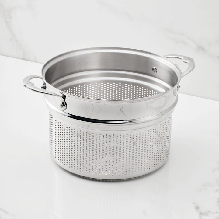 The Hestan Provisions 8-Quart Stainless Steel Pasta Insert and matching stockpot, both with handles and small, evenly spaced holes, rest on a white marble surface.