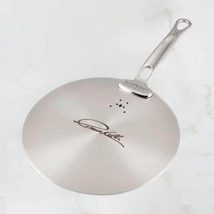 The Thomas Keller Insignia Commercial Clad Stainless Steel Universal Lid, part of our eco-friendly cookware line, boasts a smooth surface and a long handle, with an engraving on the base and three small decorative holes near the handle attachment.