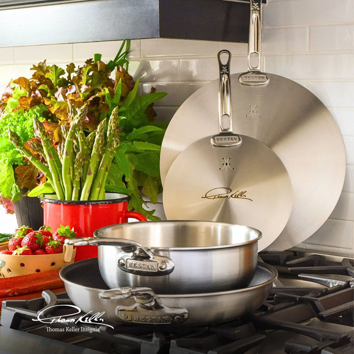 In a kitchen scene, Thomas Keller Insignias stainless steel cookware is showcased on the stove, with two pans stacked and a Commercial Clad Stainless Steel Universal Lid beside them. Two larger lids hang above, and fresh produce is presented in an eco-friendly red pot on the counter.