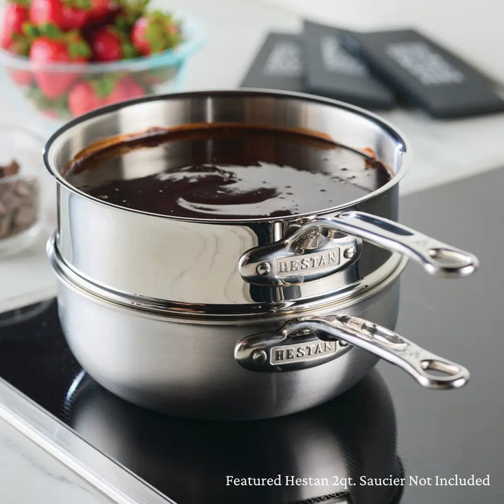 Two stacked Hestan Provisions stainless steel sauciers on an induction cooktop showcase even heat distribution. The top acts as a double boiler with melted chocolate inside. Blurred in the background are fresh strawberries, chocolate chips, and black booklets. Text: Featured 3qt Double Boiler Insert Not Included.