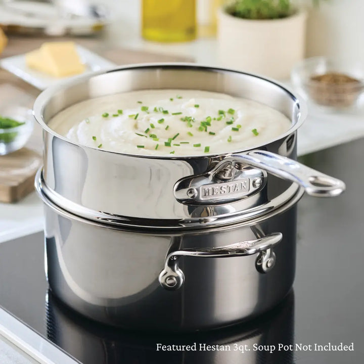 A stainless steel double boiler insert sits on a stove, filled with creamy soup topped with chopped chives. Thanks to its even heat distribution, a small saucepan holds an unopened package of butter in the background. The pot proudly displays the brand name Hestan Provisions on its handle.