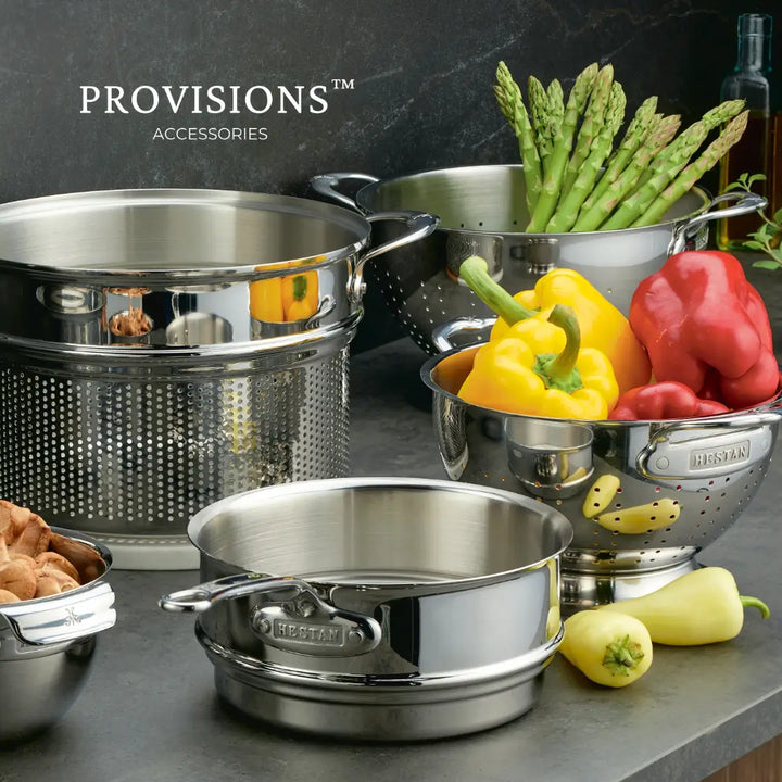 A kitchen scene showcases Hestan Provisions stainless steel double boiler inserts known for even heat distribution and colanders filled with asparagus, bell peppers, and zucchini. A small pot with mushrooms is visible, branded as Hestan Provisions.