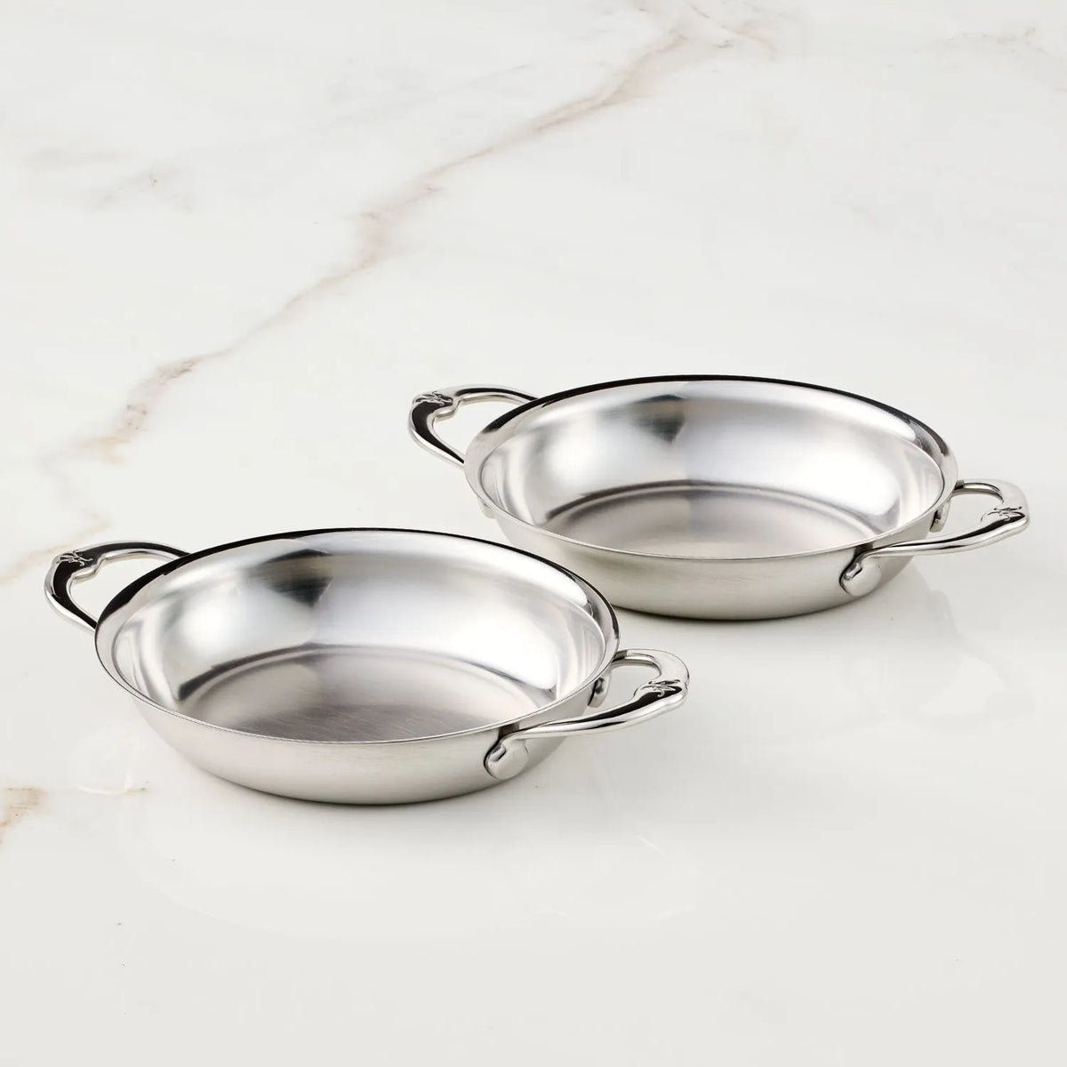 Two Hestan Provisions 6.5"x5.5" oval au gratin dishes (2-piece set) with side handles are displayed on a white marble surface.
