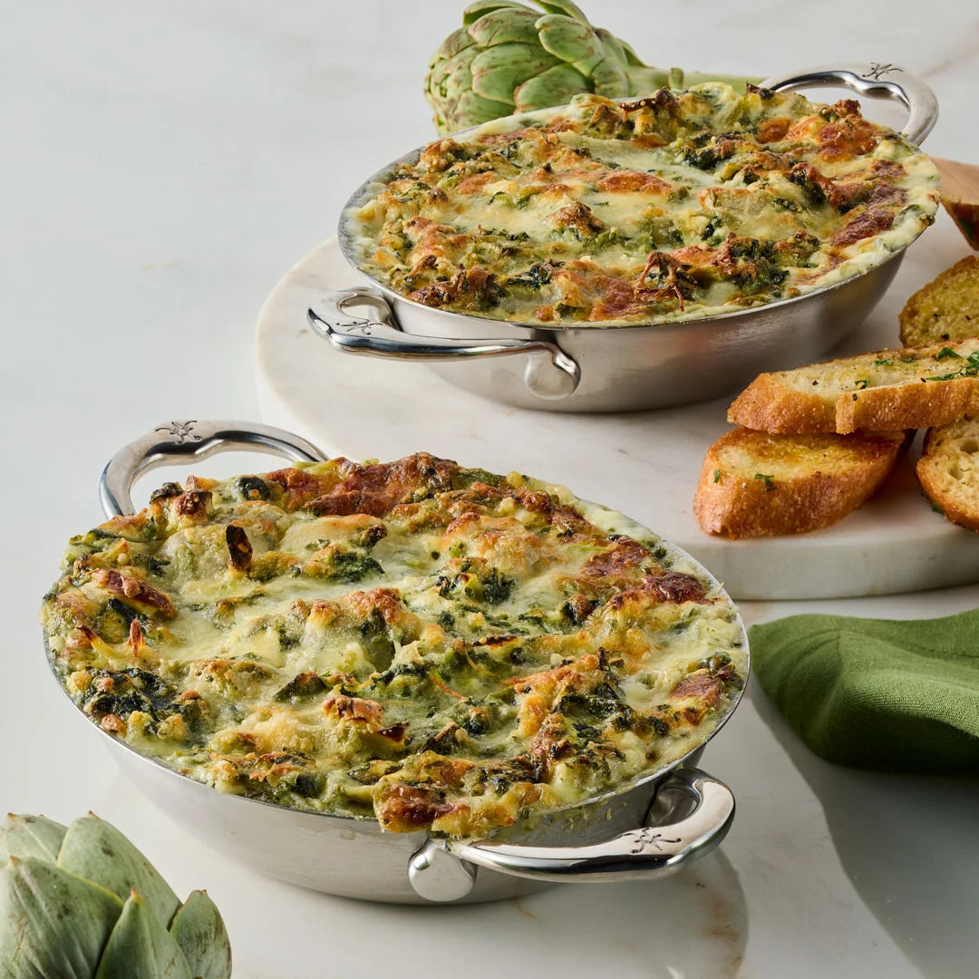 Two Hestan Provisions 6.5-Inch x 5.5-Inch Oval Au Gratin dishes are filled with cheesy spinach artichoke dip on a marble surface, accompanied by sliced toasted bread, artichokes, and a green napkin. Set includes two pieces.