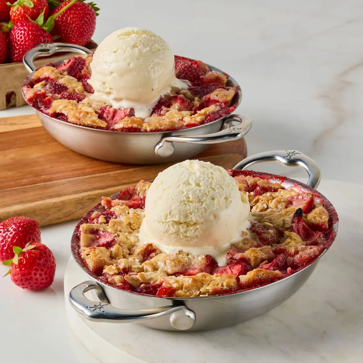 Two small stainless steel Hestan Provisions au gratin pans, each with a scoop of vanilla ice cream atop strawberry crumble, rest on marble. Fresh strawberries lie on a wooden board in the background, resonating with Michelin-starred chefs appreciation for the Small Oval Au Gratin Set.