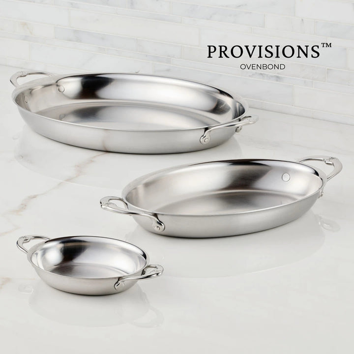 The Small Oval Au Gratin Set by Hestan Provisions, featuring two stainless steel baking dishes, is displayed on a light marble surface. Favored by Michelin-starred chefs, the dishes have handles on each end. PROVISIONS™ OVENBOND appears in the background.