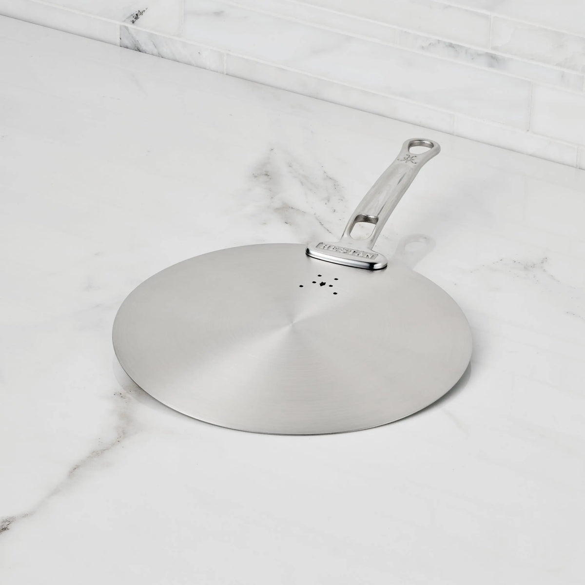 The Hestan Provisions Stainless Steel Universal Lid, featuring a handle and splatter screen, rests on a white marble countertop by a tiled backsplash; its dishwasher safe for an eco-friendly kitchen.