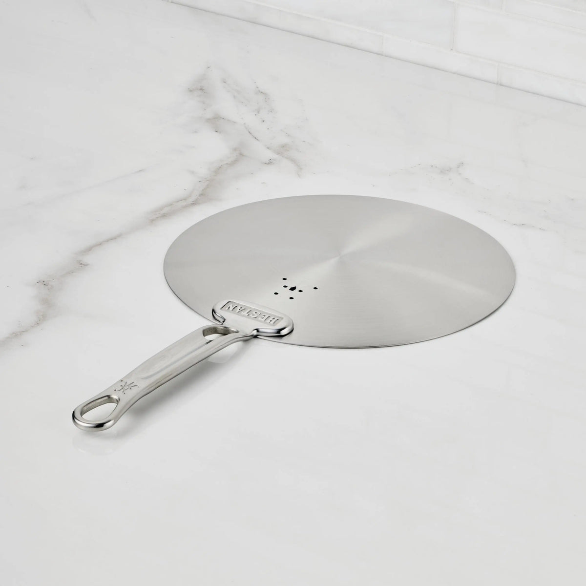 A Hestan Provisions Provisions Stainless Steel Universal Lid with a handle sits on a white marble counter. Featuring three center holes, it is dishwasher safe, and “MESSER” is engraved on the handle.