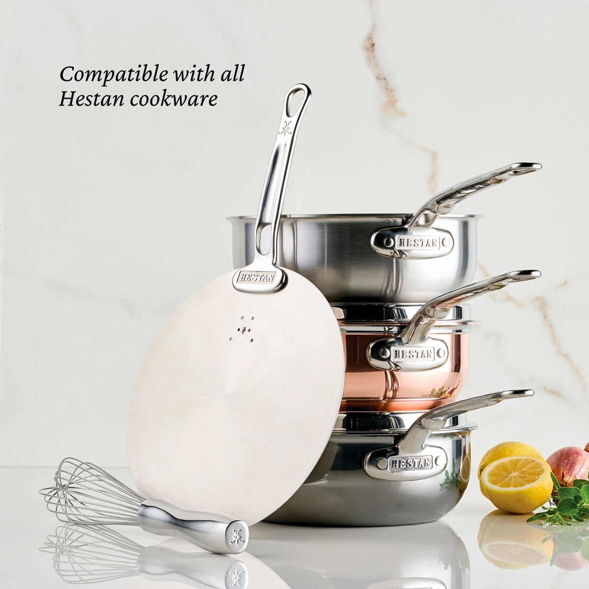 Three stacked Hestan Provisions saucepans in silver and copper with a Provisions Stainless Steel Universal Lid sit next to a whisk, lemon halves, and parsley on marble; text reads Compatible with all Hestan Provisions cookware.