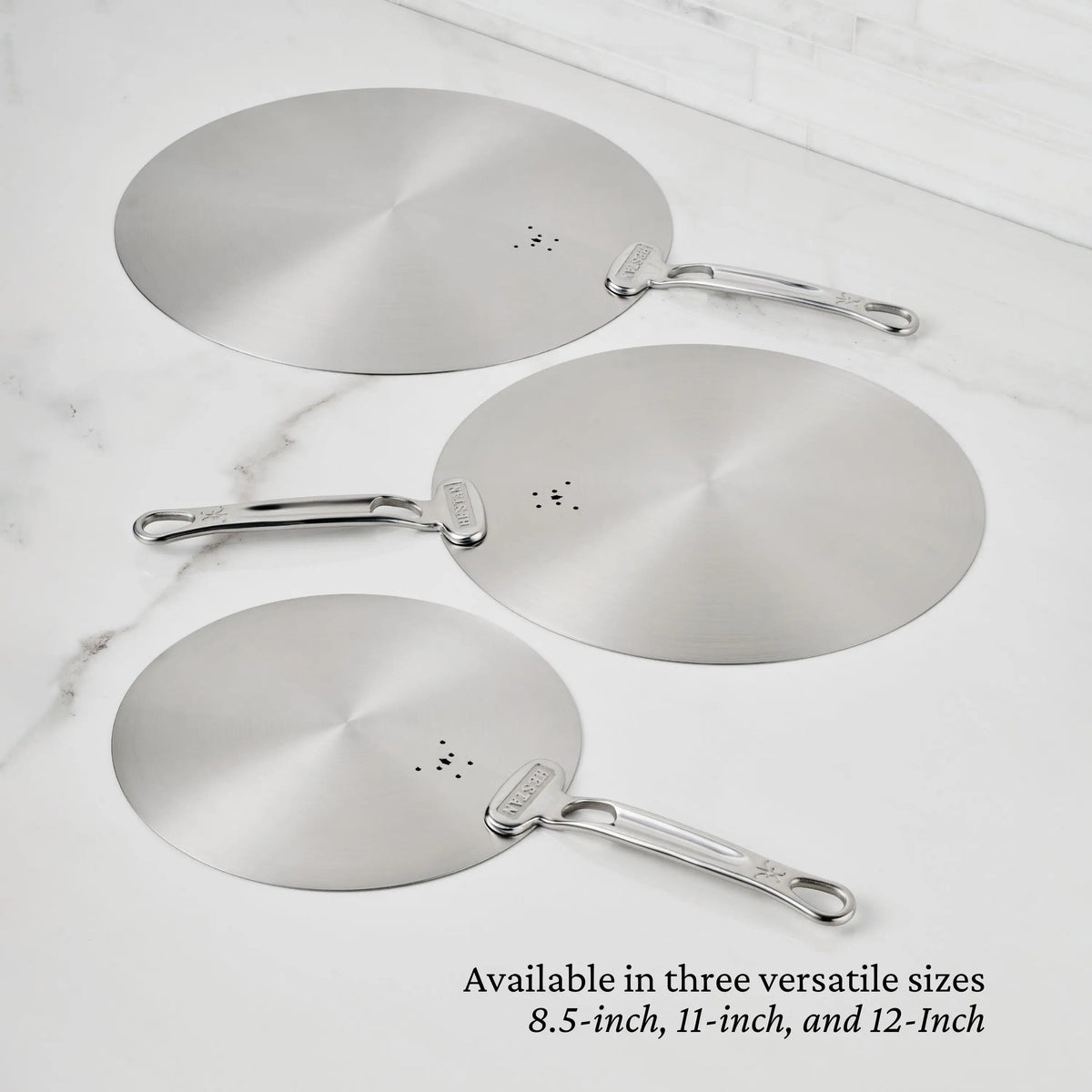 Three Hestan Provisions Stainless Steel Universal Lids with handles are shown on a white marble surface. These eco-friendly lids come in 8.5-inch, 11-inch, and 12-inch sizes and are dishwasher safe for easy cleaning.