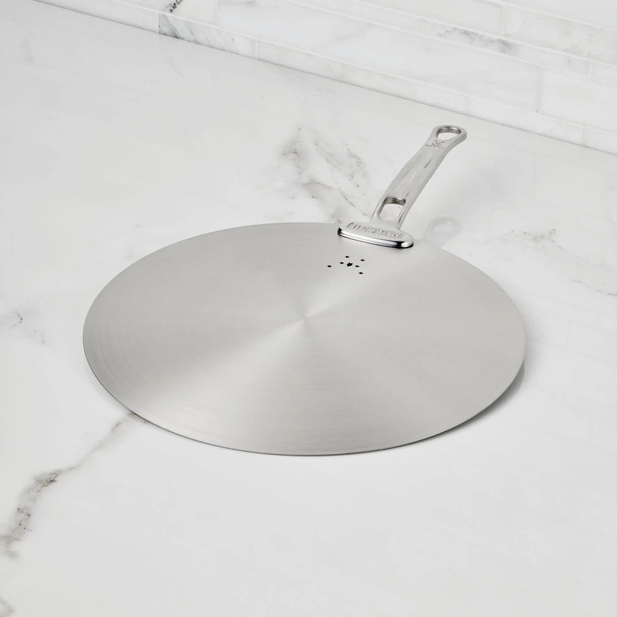 The Hestan Provisions Stainless Steel Universal Lid serves as both a splatter screen and lid, features a handle, rests on a white marble countertop with gray veining, is eco-friendly, and dishwasher safe—an essential for any kitchen.