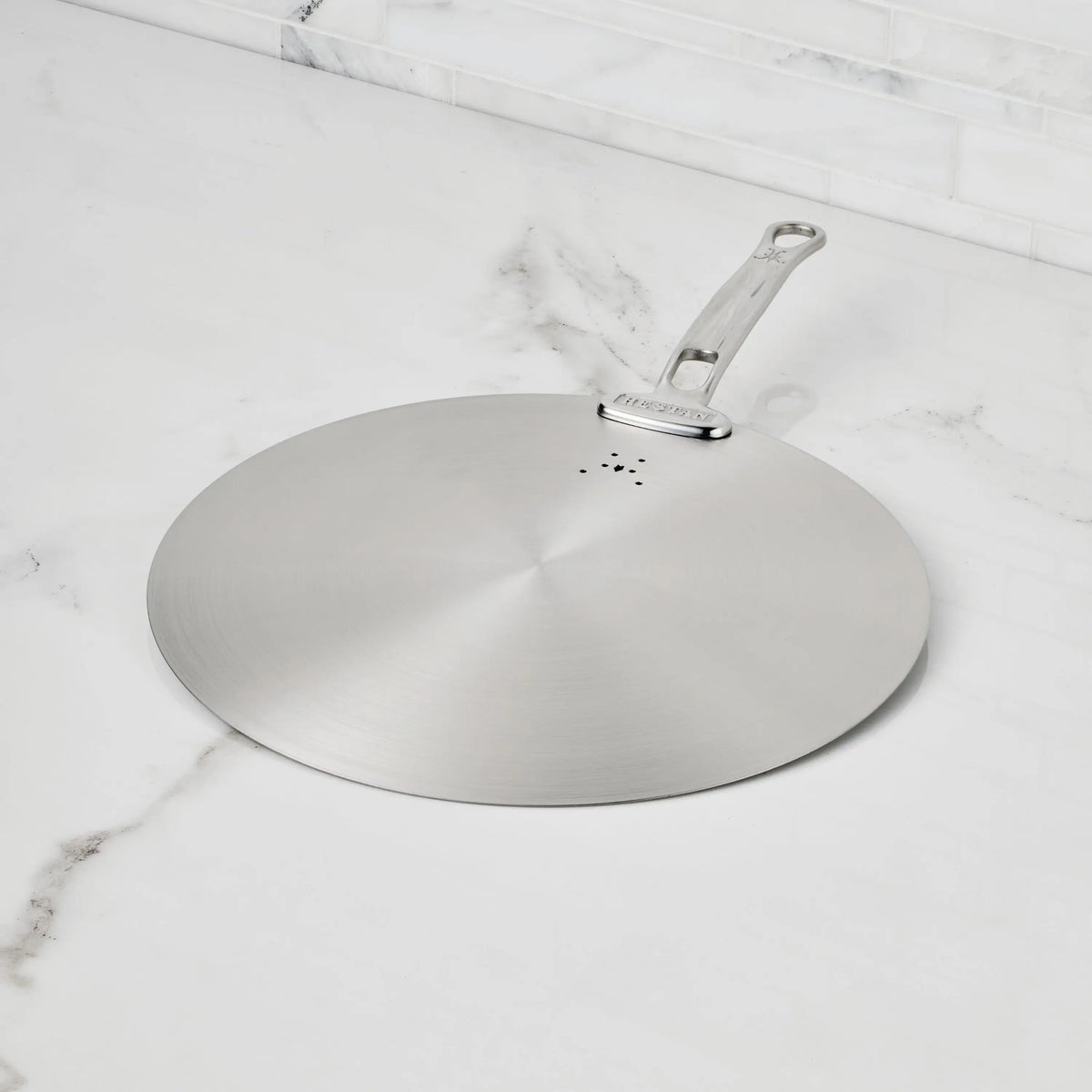 The Hestan Provisions Stainless Steel Universal Lid serves as both a splatter screen and lid, features a handle, rests on a white marble countertop with gray veining, is eco-friendly, and dishwasher safe—an essential for any kitchen.