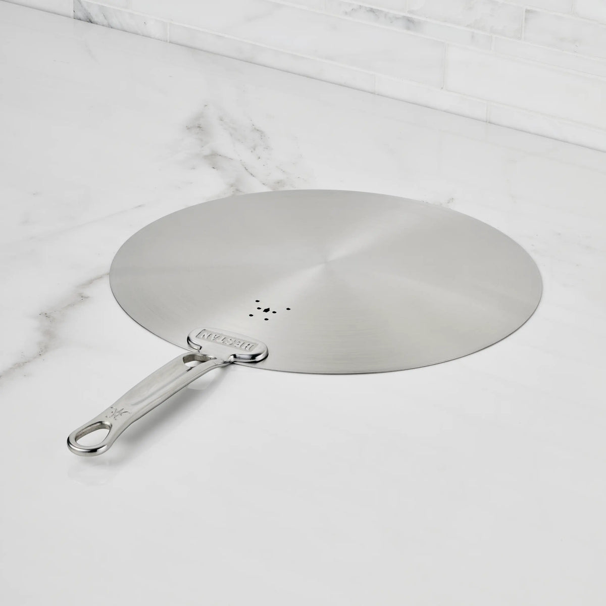 The Hestan Provisions Stainless Steel Universal Lid rests flat on a white marble countertop, blending into your eco-friendly kitchen with its dishwasher-safe design and built-in splatter screen.