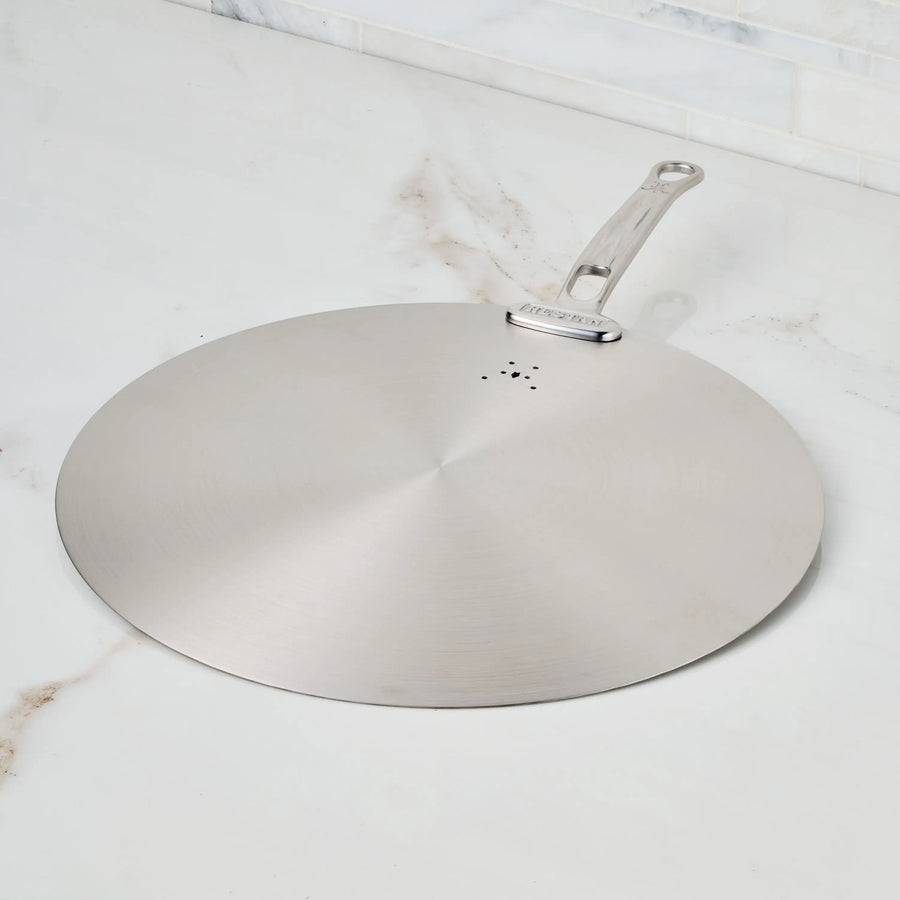 The Hestan Provisions Stainless Steel Universal Lid, a dishwasher-safe splatter screen with a metal handle and small ventilation holes, rests on a white marble countertop—perfect for any eco-friendly kitchen.