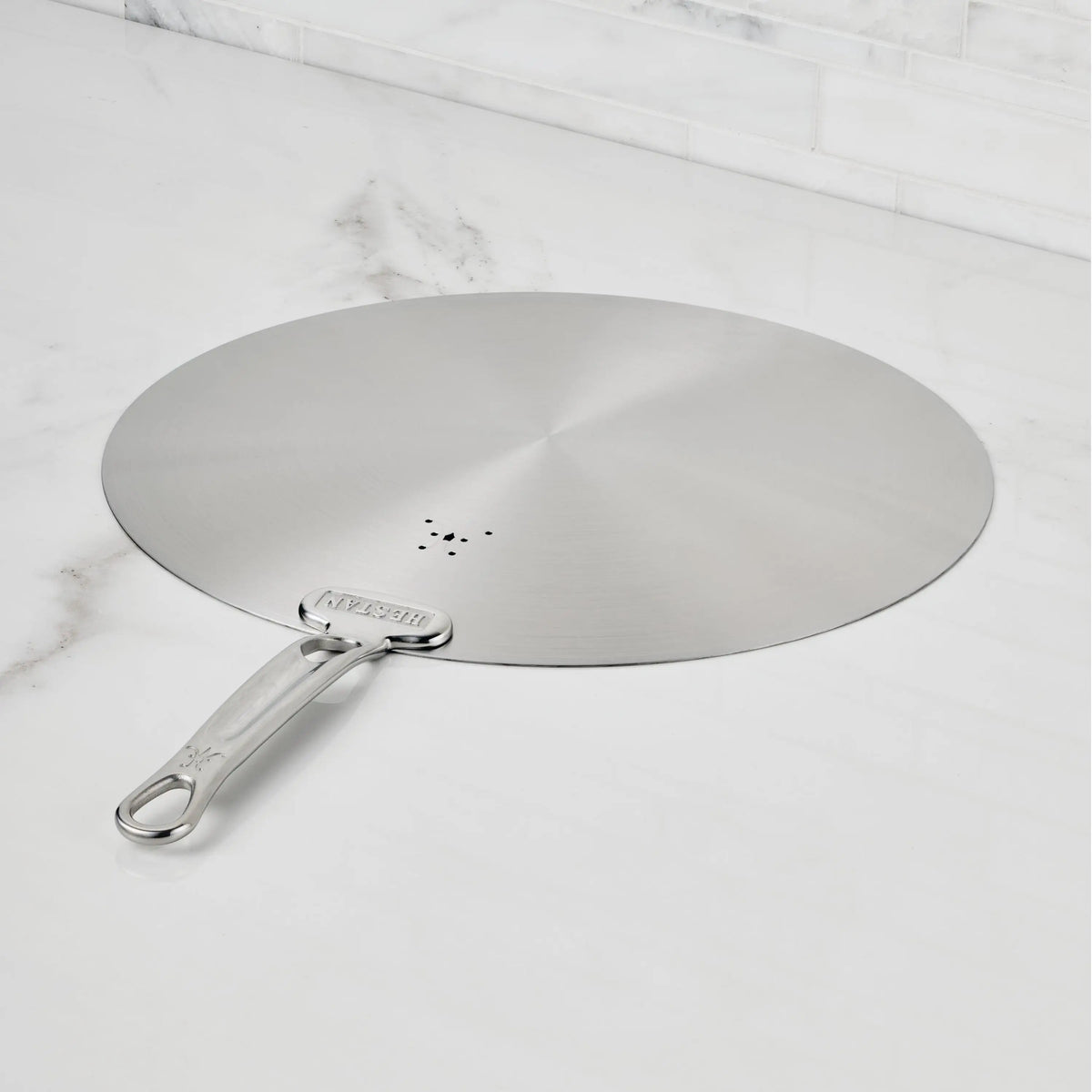 The Hestan Provisions Stainless Steel Universal Lids feature a long handle, central small holes, and can serve as a splatter screen—an eco-friendly, dishwasher safe kitchen essential displayed on a white marble countertop.