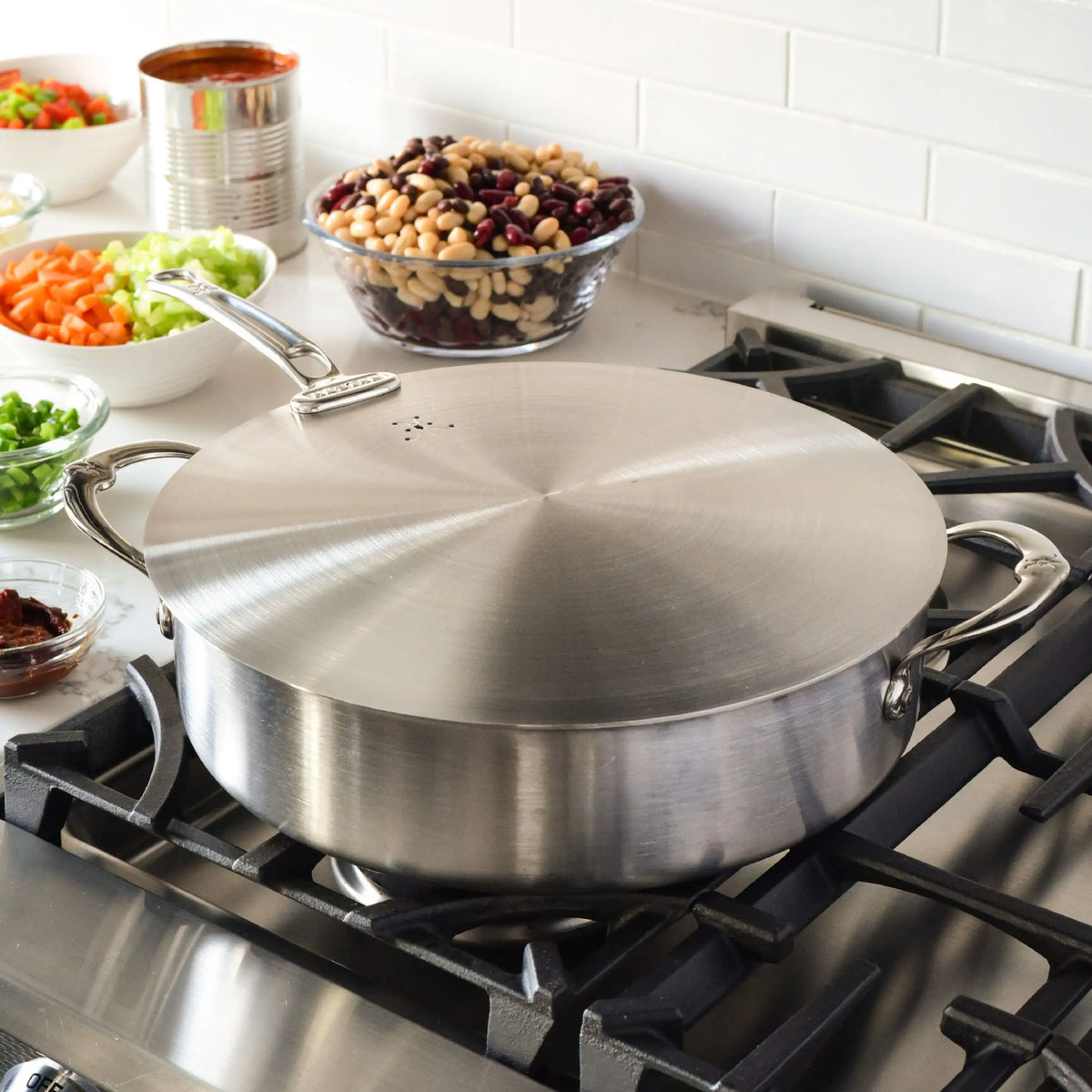 A large stainless steel pot with a Hestan Provisions Stainless Steel Universal Lid sits on a stove, while bowls of chopped vegetables, beans, and a can of tomato sauce rest on an eco-friendly counter with a white tile backsplash.