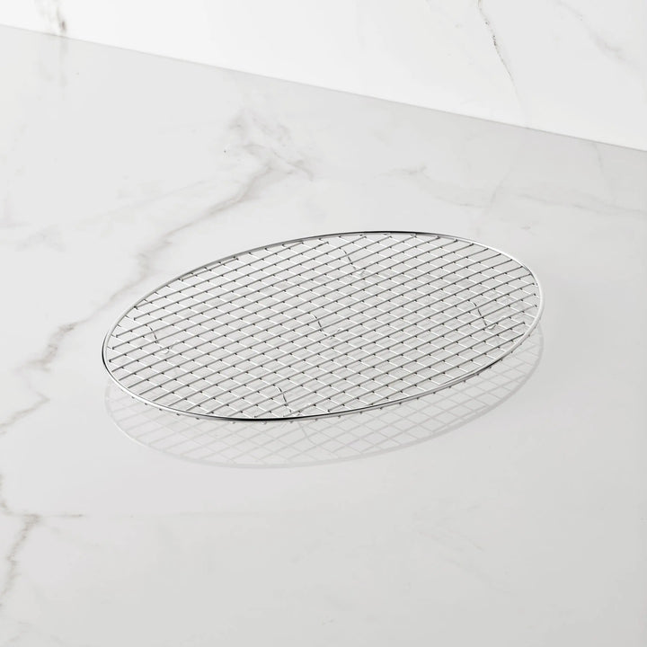 A Hestan Provisions 17.5-Inch x 14-Inch Clad Oval Roaster & Rack rests on a white marble surface. The stainless steel rack features a grid pattern and lies flat, casting a gentle shadow beneath it.