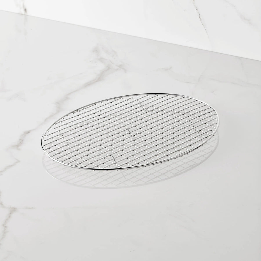 A Hestan Provisions 17.5-Inch x 14-Inch Clad Oval Roaster & Rack rests on a white marble surface. The stainless steel rack features a grid pattern and lies flat, casting a gentle shadow beneath it.