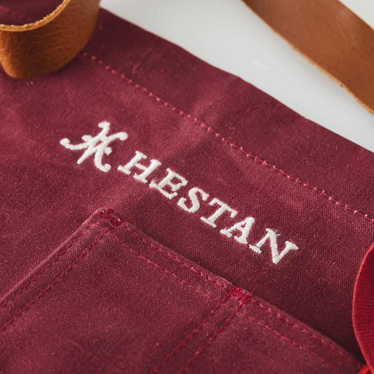 Close-up of the maroon Titanium Grilling Essentials Cookware Set, 6-piece by NanoBond, featuring HESTAN and a decorative H logo embroidered in white above a pocket; a brown leather strap is partially visible at the top.