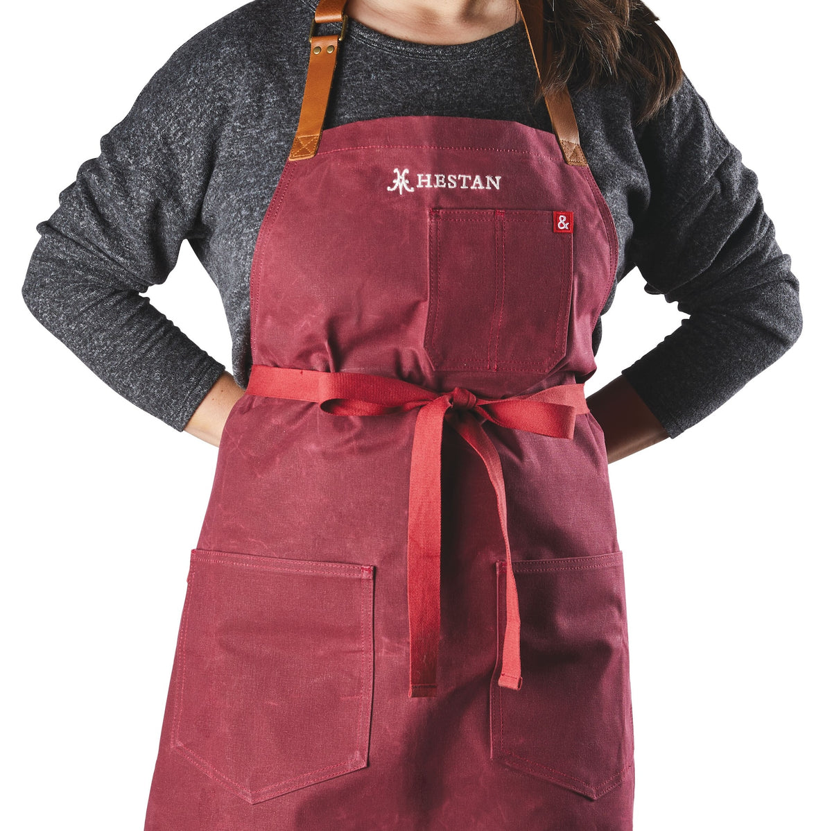 A person wearing a dark gray long-sleeve shirt and a red premium grilling apron with two front pockets, leather neck strap, front tie, and HESTAN embroidery is using the NanoBond Titanium Grilling Essentials Cookware Set (6-piece).