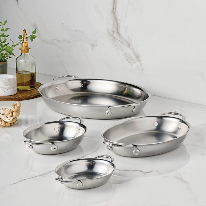 Four Hestan Provisions 8.5-Inch x 6.5-Inch Oval Au Gratin pans with handles are displayed on a white marble countertop, with a bottle of oil, a small plant, and wooden beads arranged on a round tray in the background.