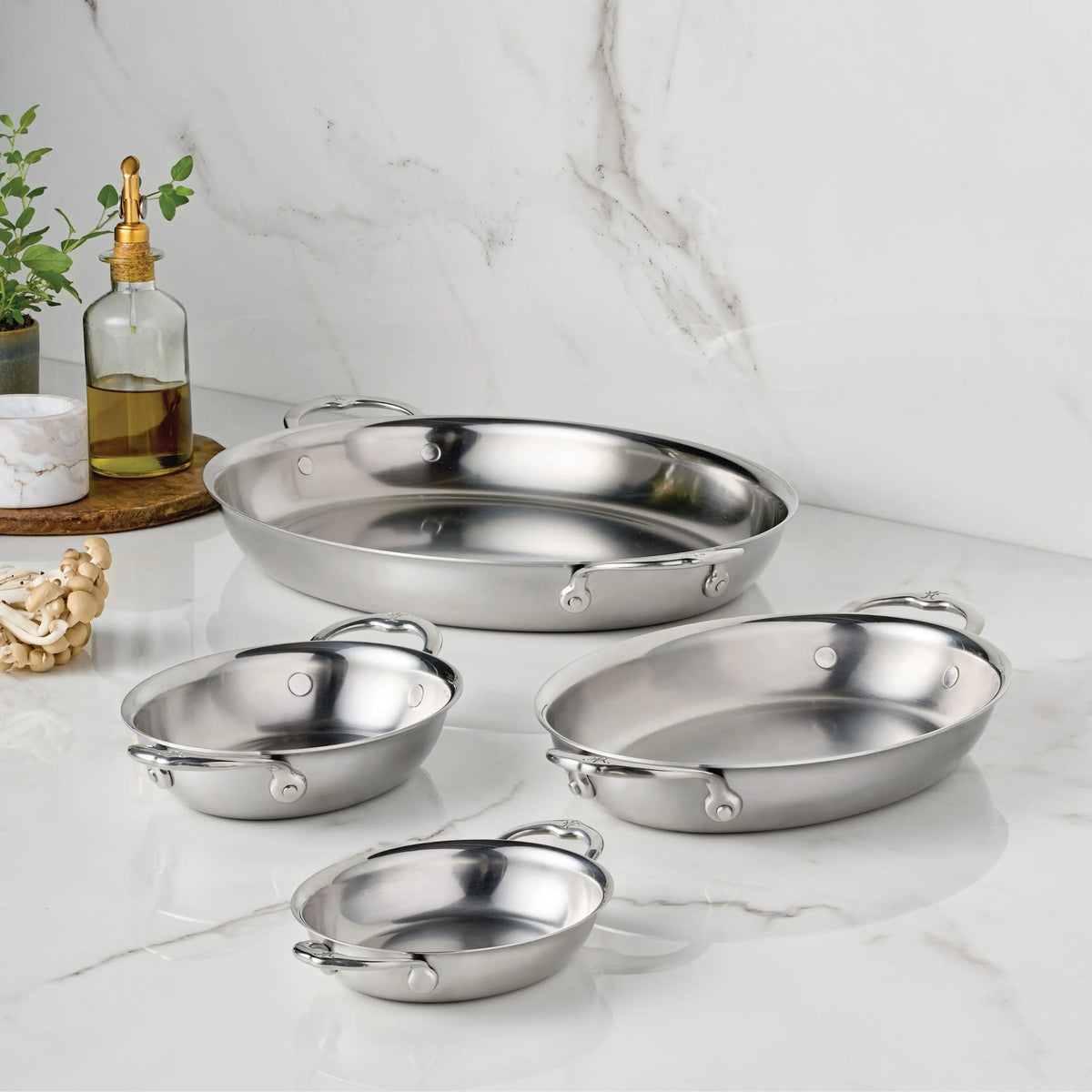 Four Hestan Provisions 8.5-Inch x 6.5-Inch Oval Au Gratin pans with handles are displayed on a white marble countertop, with a bottle of oil, a small plant, and wooden beads arranged on a round tray in the background.