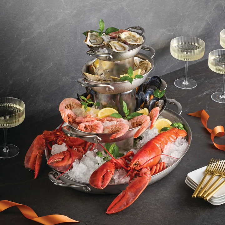 A three-tiered seafood tower with oysters, clams, shrimp, mussels, and lobsters rests on ice. Nearby are lemon wedges, mint, white wine glasses, gold forks, and a gleaming Hestan Provisions 21" x 14" Clad Oval Roaster.