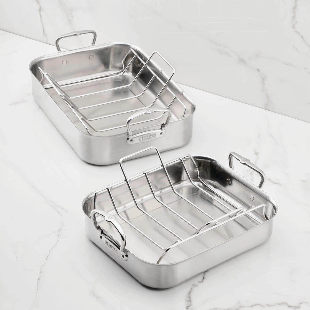 Two empty Hestan Provisions Brushed Clad Stainless Steel Roasters with racks and sturdy side handles sit on a flawless white marble surface.