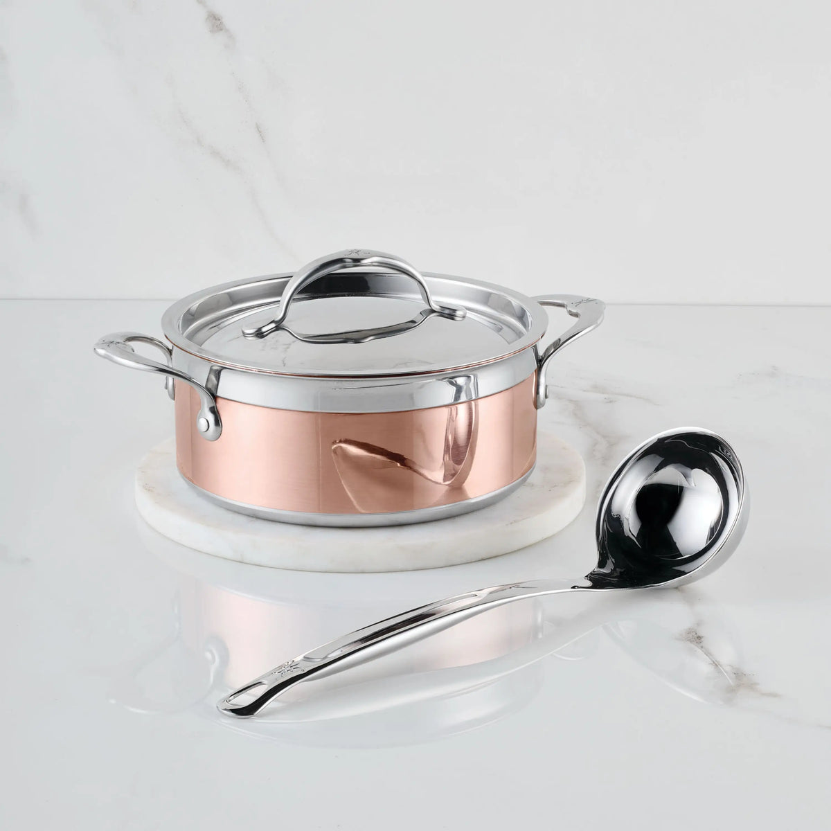 A CopperBond Copper Induction Soup Set, 3-Piece, sits on a round white marble surface with a large metal ladle in front. The handcrafted pot and lid are set against a clean white marble background.