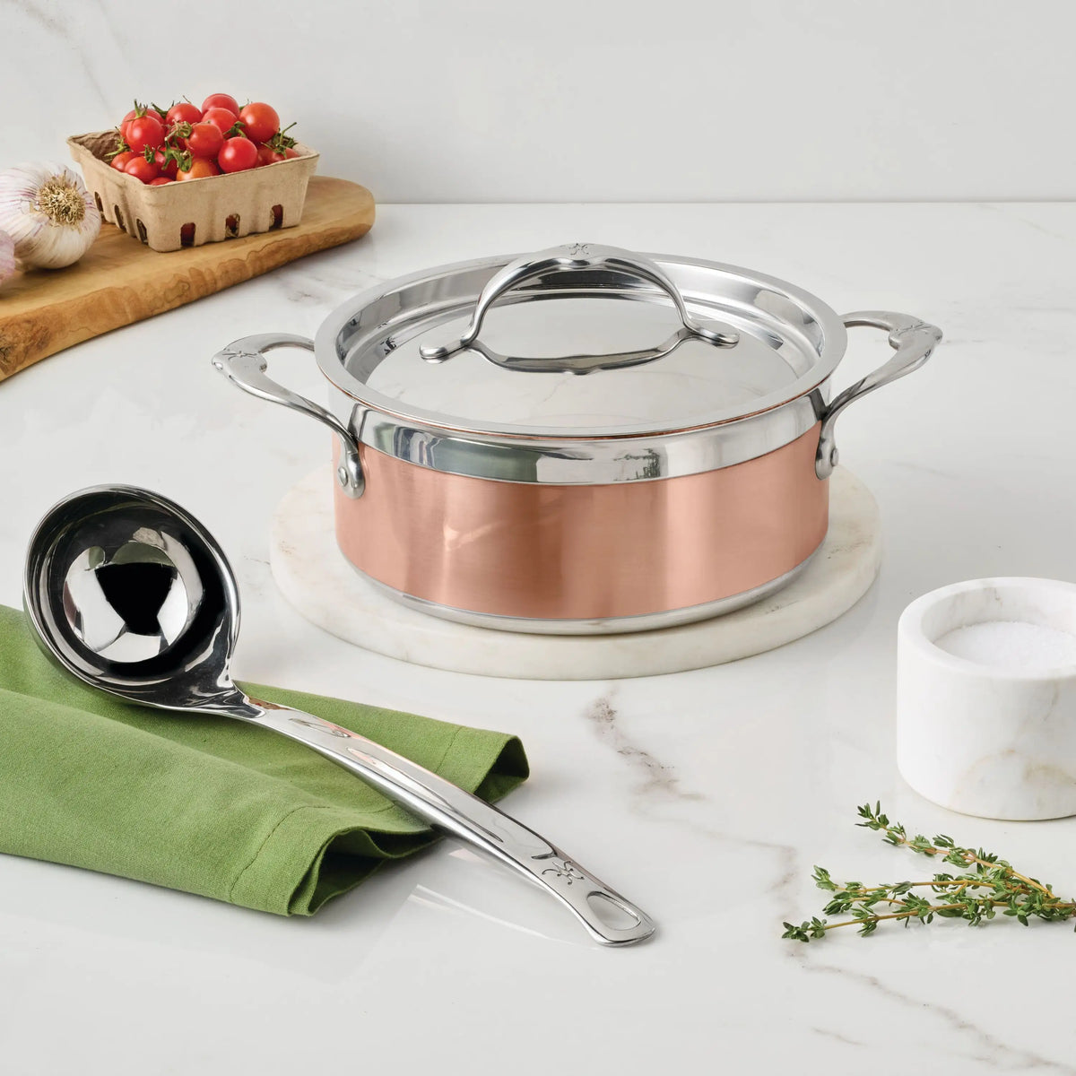 A CopperBond Copper Induction Soup Set, 3-piece, handcrafted in Italy with lid, rests on marble. Nearby are a stainless steel ladle on a green cloth, a marble salt cellar, fresh thyme, and a wooden board with tomatoes and garlic.