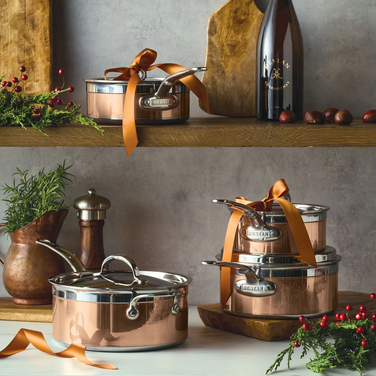 A kitchen scene features CopperBond Copper Induction Saucepans and other copper pots with lids, some tied with orange ribbons, plus a wine bottle, wooden cutting board, pepper grinder, ceramic jug, and festive greenery on wooden shelves.