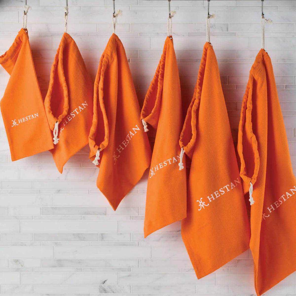 Six orange Accessories Flannel Cookware Storage Bags with white logos hang evenly on hooks against a white tile wall, each featuring a rope loop for easy hanging.