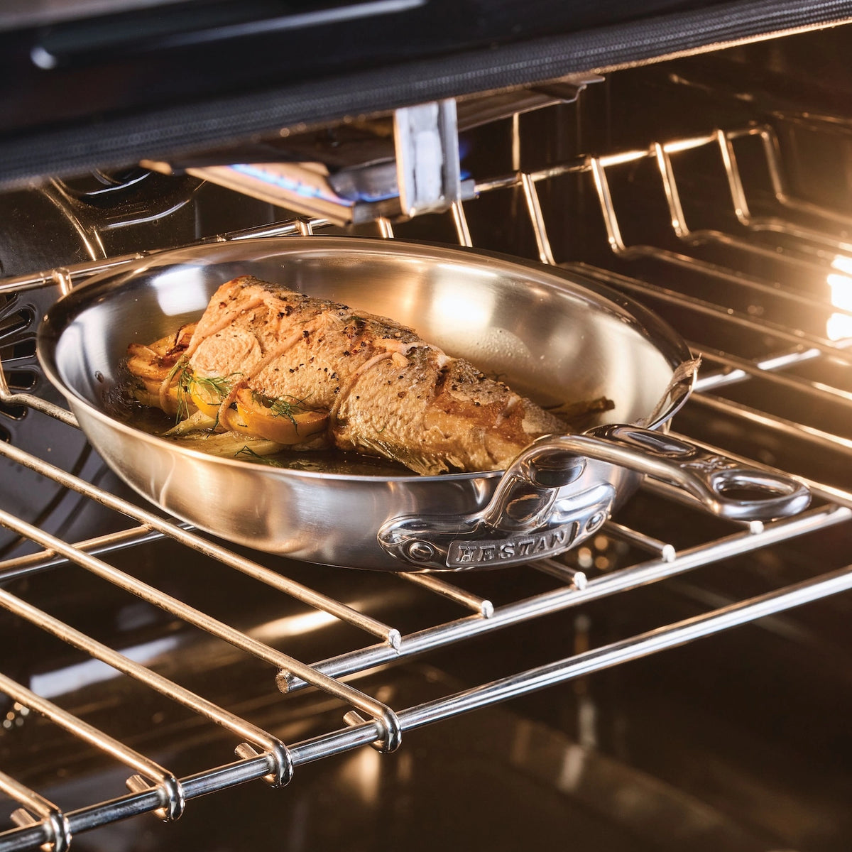 A Hestan Provisions Oval Fish Pan sizzles with seasoned salmon on a wire rack in the oven, ensuring even heat distribution, while the light highlights the garnished fish.