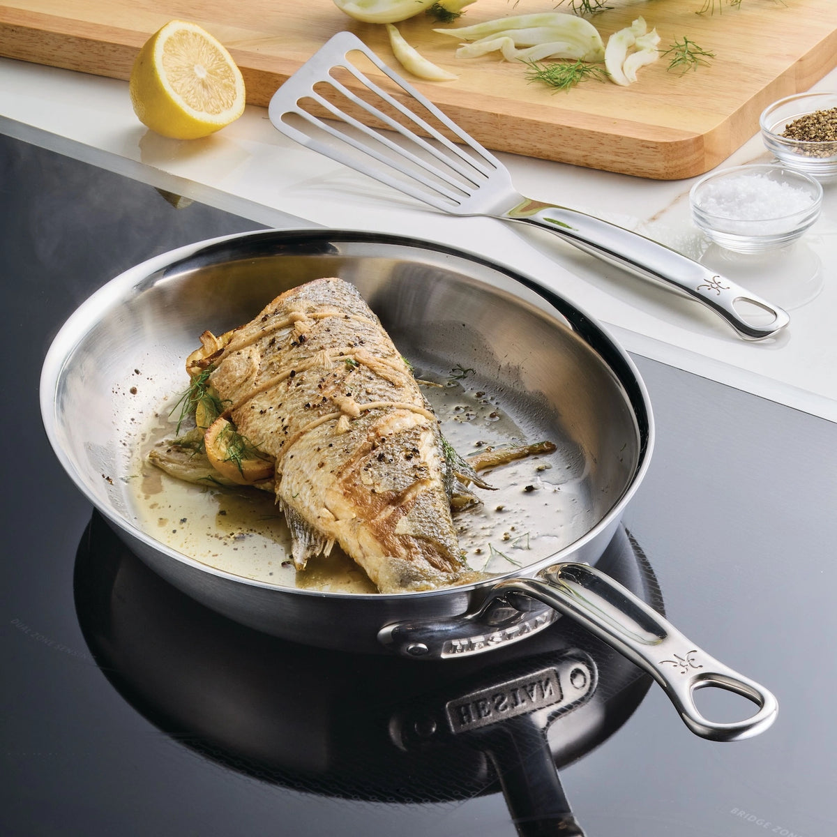 A fish with herbs sizzles in a Hestan Provisions Oval Fish Pan for even stovetop heat. Nearby, a spatula lies next to sliced fennel, half a lemon, salt, and pepper on a cutting board. Steam rises from the pan.