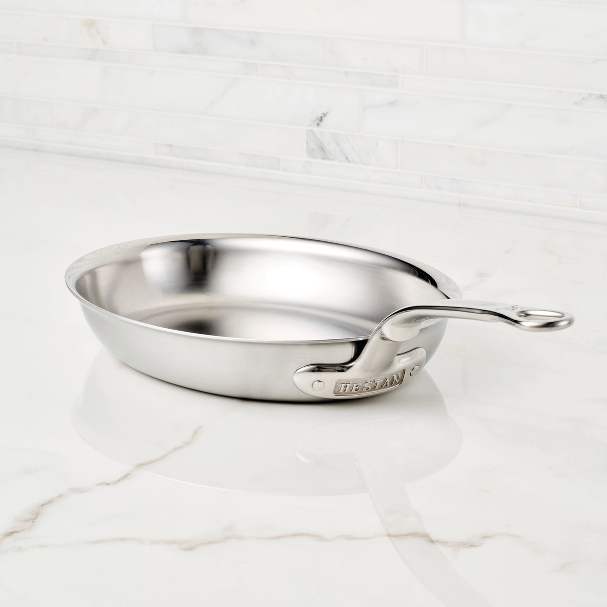 A stainless steel Hestan Provisions Oval Fish Pan, renowned for even heat distribution, shines on a white marble countertop. Its sturdy handle is engraved with the brands name, and a white tiled wall enhances the sleek kitchen backdrop.