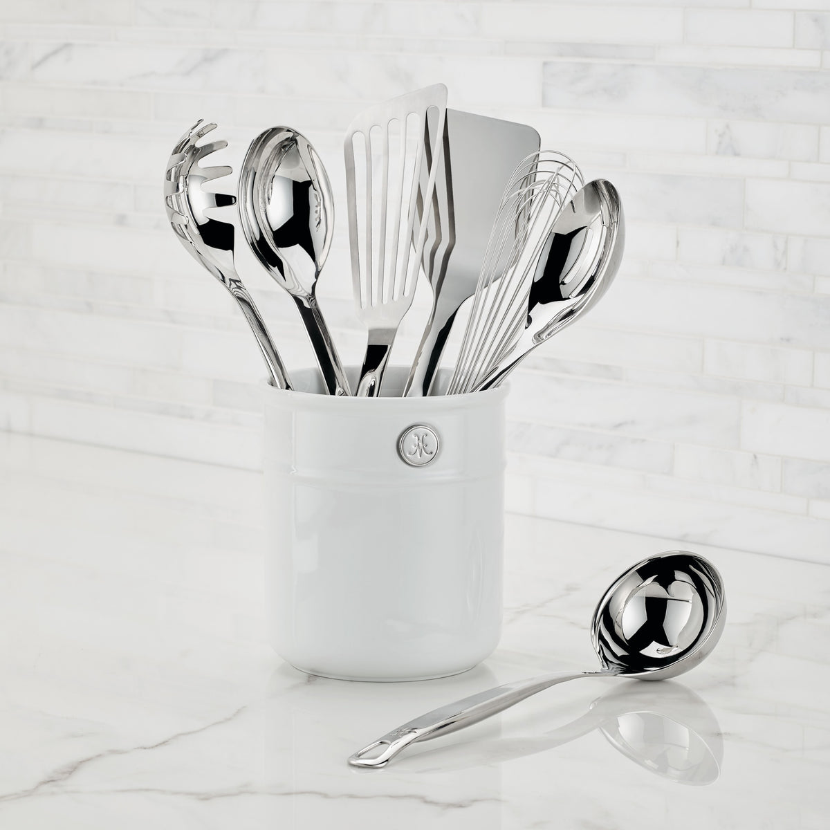 A white ceramic utensil holder showcases the Hestan Provisions OvenBond Tri-ply Epicurean Stainless Steel 21-Piece Set of kitchen tools like spoons, spatulas, and a whisk against a white tiled backsplash, with a ladle elegantly placed on the countertop.