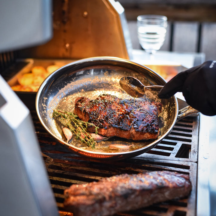 A gloved hand spoons sauce over a seared steak with herbs and garlic in the NanoBond Titanium x Tyler Florence 41-Piece Cookware Set on an outdoor grill. Also visible are meat, potatoes, and a glass of water in the background.