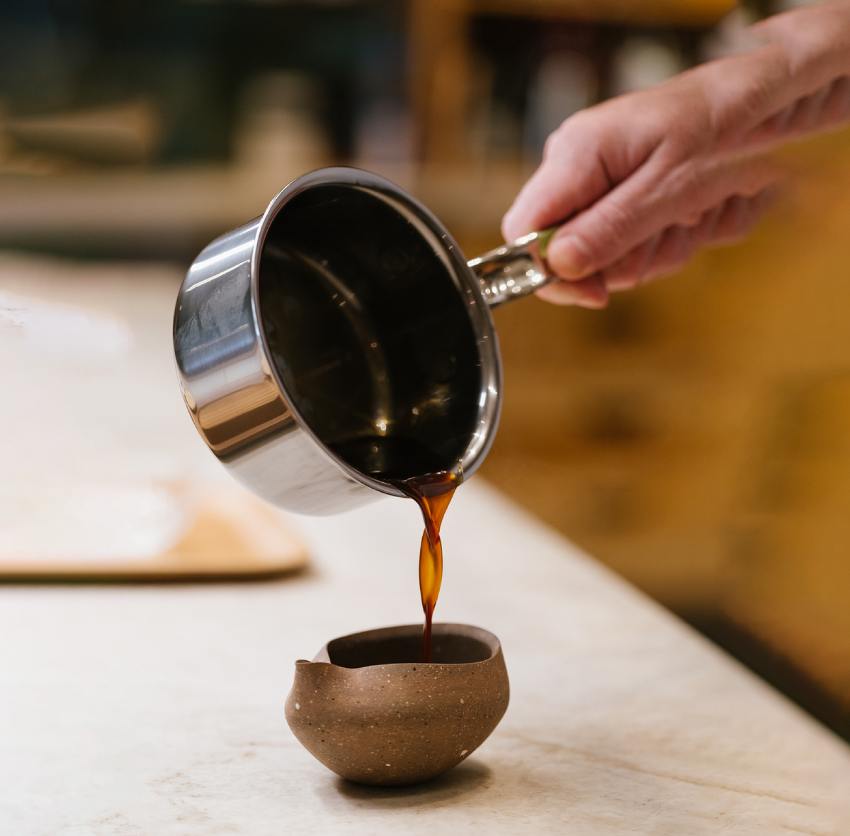 A hand pours dark liquid from a NanoBond saucepan, part of the 18-Piece Titanium Cookware Set with Dutch Oven, into a small brown ceramic cup on a light countertop.