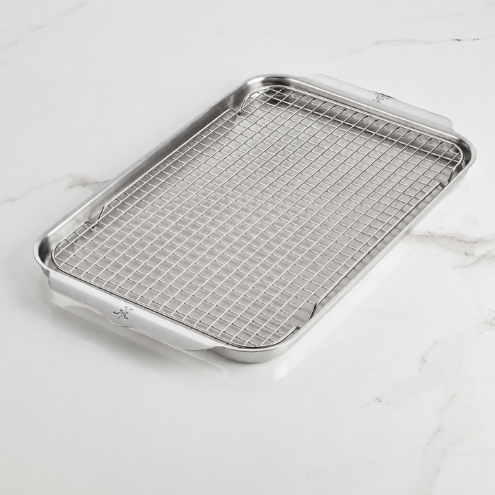 The Hestan OvenBond Tri-ply Half Sheet Pan & Rack, 13-in x 18-in, is a rectangular stainless steel pan with raised edges and a removable metal rack, shown on a white marble surface.