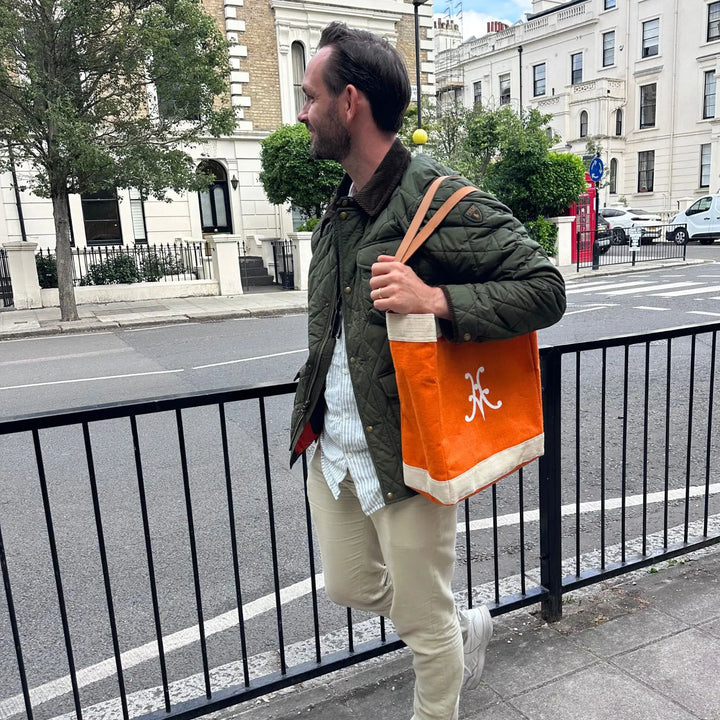 A man stands on a sidewalk by a black railing, wearing a green quilted jacket, light pants, and white sneakers. He carries the Accessories Hestan Market Tote in Citra Orange. Urban buildings and a parked car are in the background.