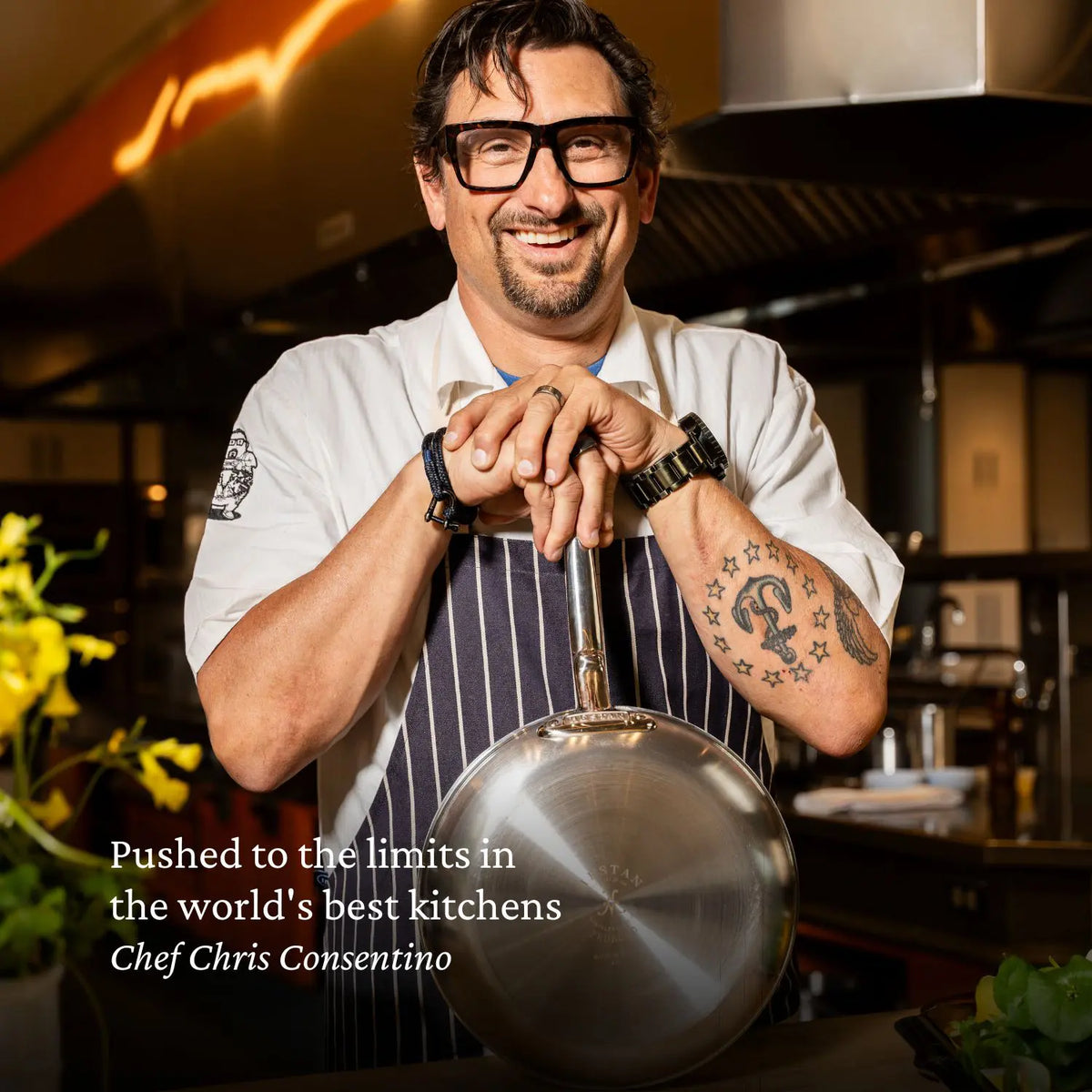 A man in glasses and a white chef’s coat stands smiling in a pro kitchen, holding a Brushed Clad Stainless Steel Saucepan with Lid by ProBond® Forged™. Text reads: Pushed to the limits—handcrafted in Italy, trusted by Chef Chris Cosentino.