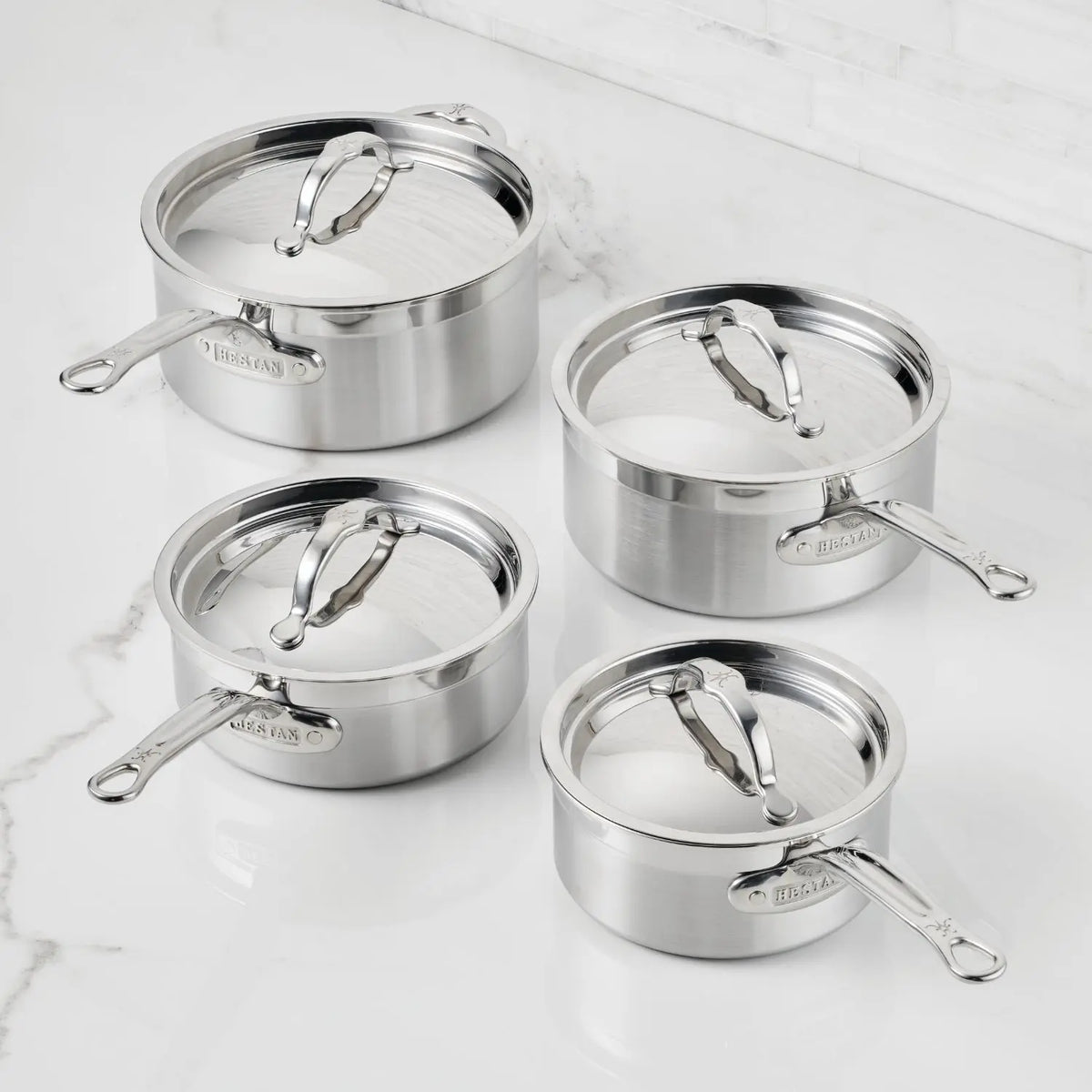 Four ProBond® Forged™ Brushed Clad Stainless Steel Saucepans, each a different size with matching lids and long handles, are arranged on a white marble countertop.