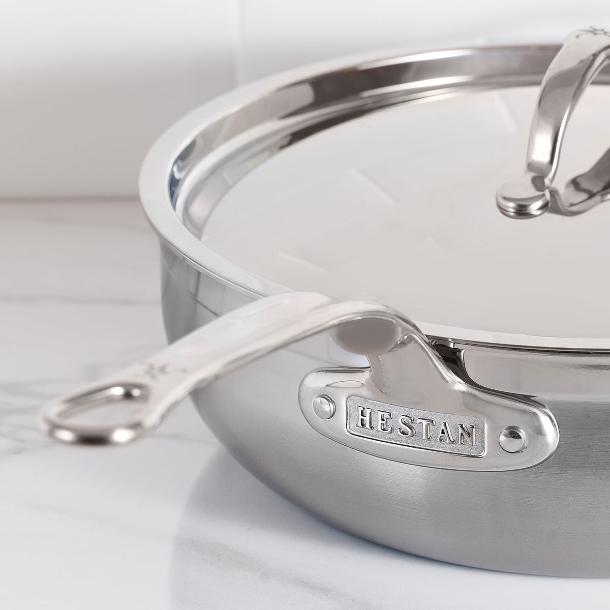 Close-up of a ProBond Professional Clad saucepan with a lid on a white countertop. The handle is in the foreground with HESTAN engraved on the tri-ply stainless steel side, and the lid has a curved top handle.